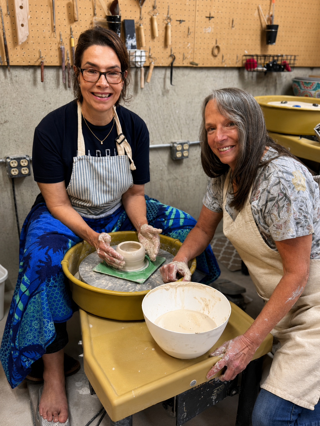 Two women laughing together at the pottery wheel in the studio